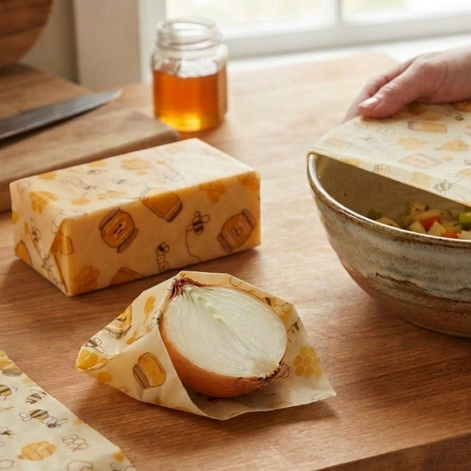 Beeswax wraps with a honeycomb and bee pattern shown in a kitchen setting, wrapping a block of cheese, a halved onion, and a ceramic bowl.