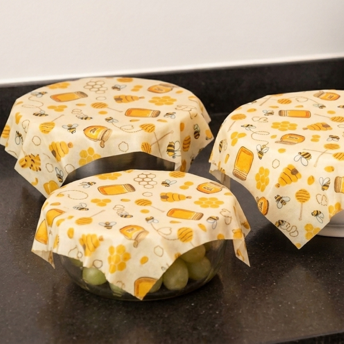Three beeswax wraps with a honeybee and honeycomb pattern in yellow and orange, shown covering glass bowls on a dark countertop.