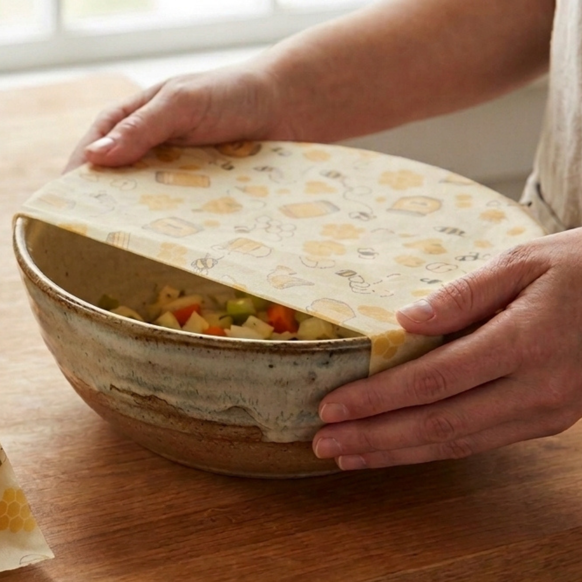 A person's hands are shown covering a bowl with a beeswax wrap on a wooden table. The cover is cream-colored with a light yellow honeycomb pattern and cartoon illustrations.